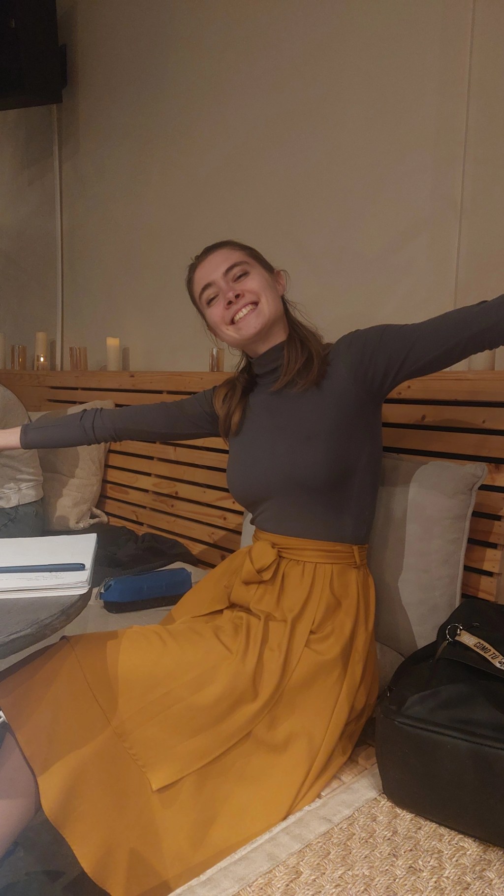Owner of the blos smiles sitting on a cozy bench in a warmly lit room, wearing a dark lilac turtleneck and mustard-yellow skirt, with arms stretched wide in a cheerful gesture.